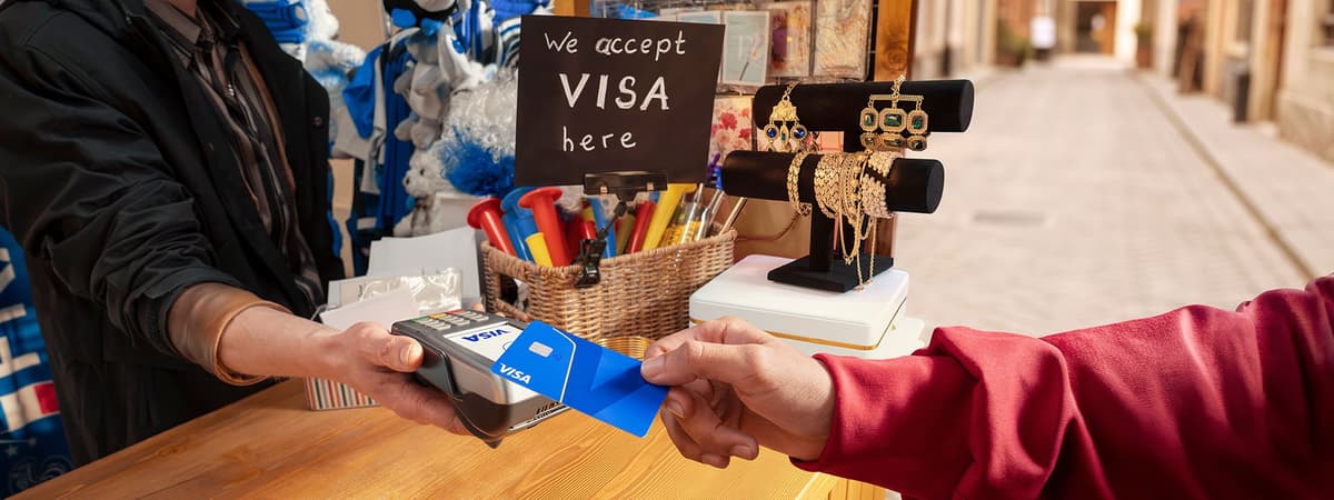 A person hands a Visa card to a shopkeeper using a card machine. A sign reads "We accept Visa here."