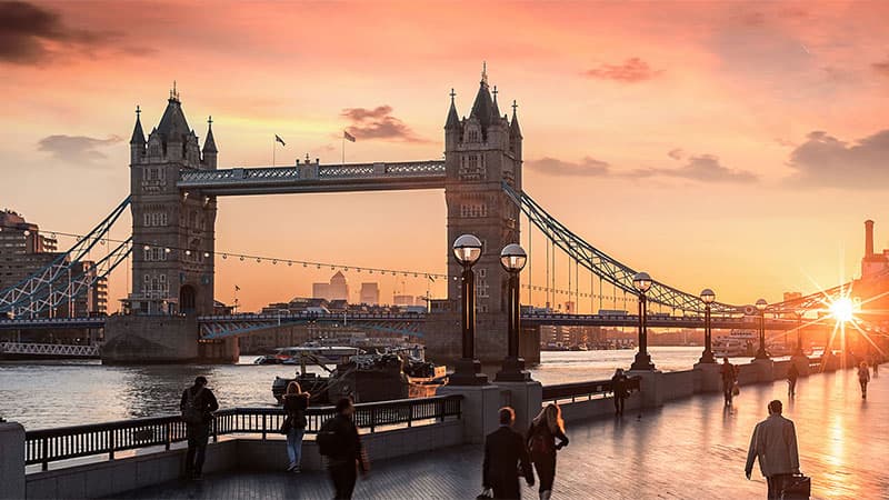 Sunset over London's Tower Bridge, with silhouettes of walking people and a setting sun reflecting on the river, creating a warm, serene ambiance.