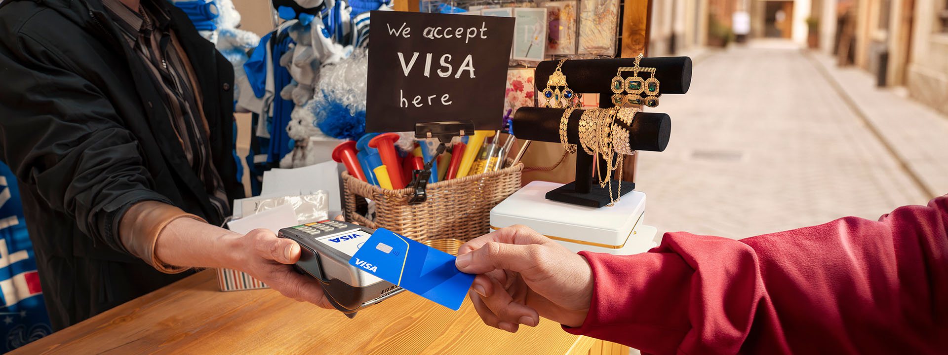 A person hands a Visa card to a shopkeeper using a card machine. A sign reads "We accept Visa here."