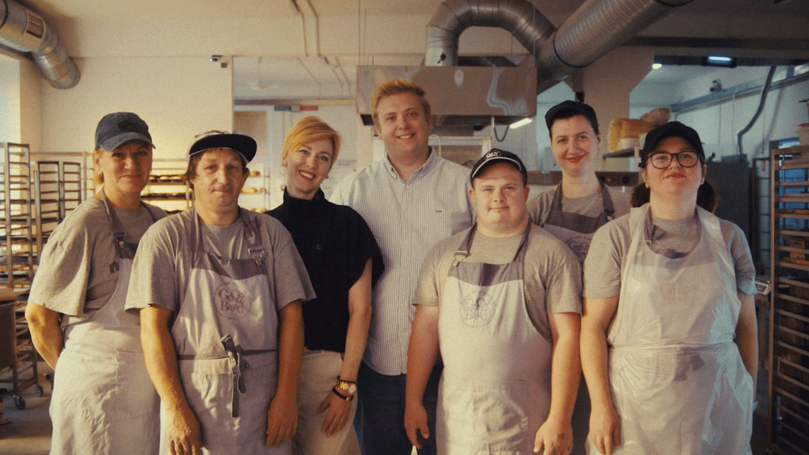 A group of seven people, some wearing aprons, pose together in a bakery setting with metal racks and industrial lighting.