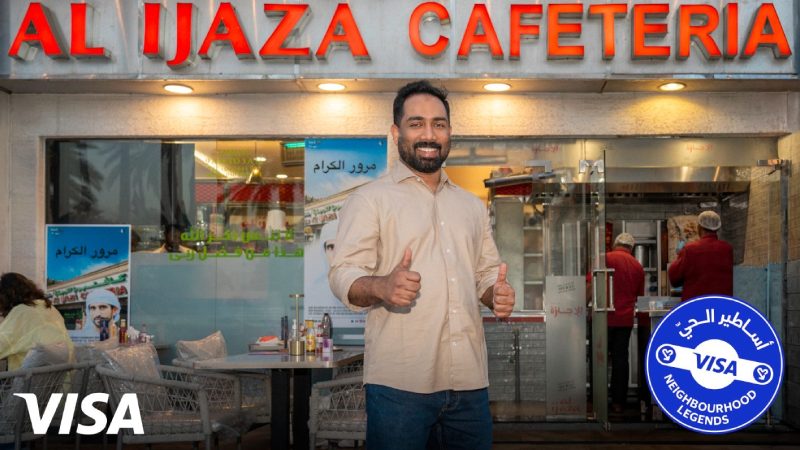 Smiling man in front of "Al Ijaza Cafeteria" with thumbs up. The scene is lively, with an inviting atmosphere. Visa logo and promotion badge visible.