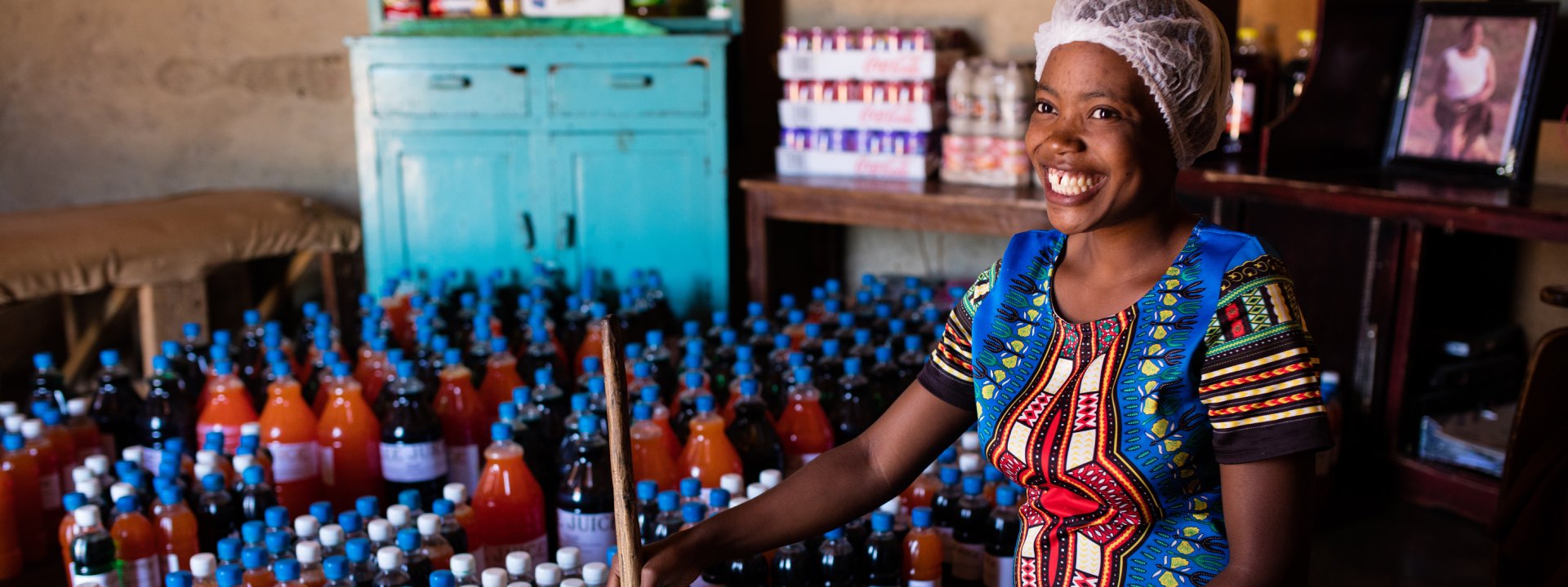 Smiling woman in colorful dress standing among bottles of homemade juice in her small business.
