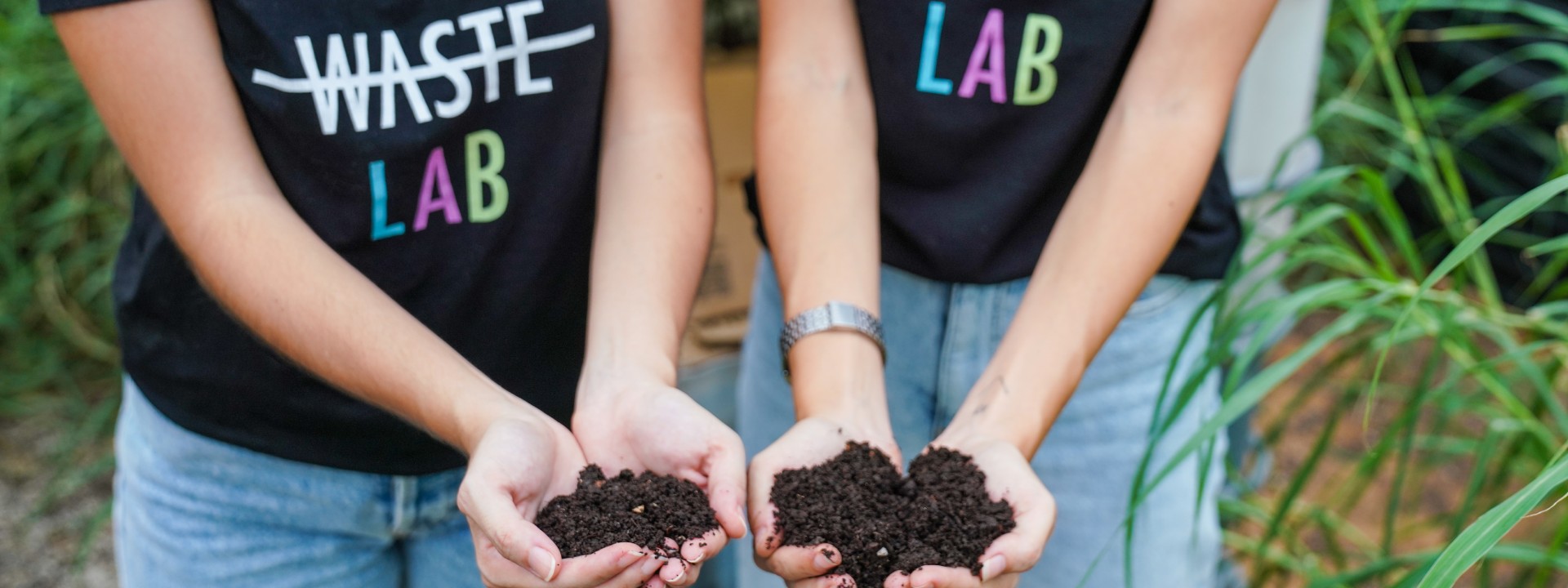 Two people holding handfuls of compost soil, wearing The Waste Lab t-shirts.