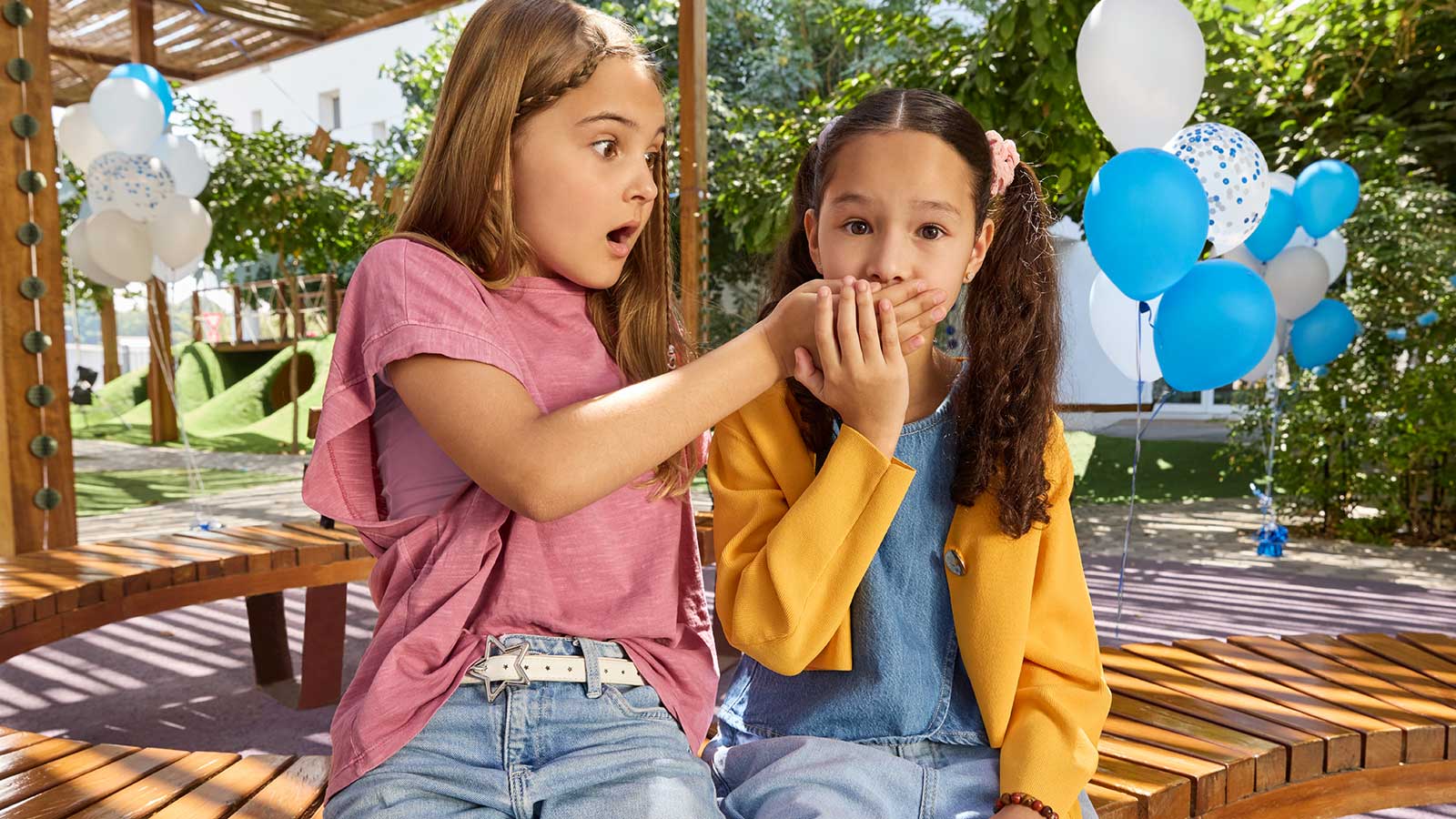 A surprised girl covers the other girl's mouth with her hand, balloons in the background