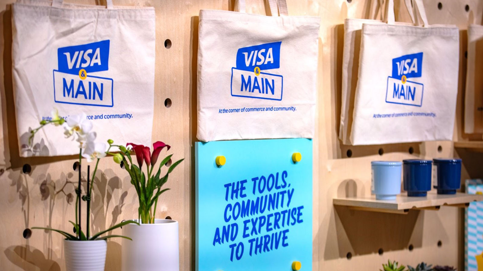 Wall with “Visa & Main” branded canvas bags hanging and a sign that says “The tools, community and expertise to thrive”